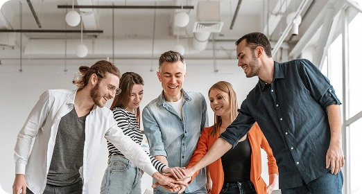 Een groep van vijf collega’s in een modern kantoor glimlacht en vouwt de handen in elkaar als teken van teamwork en saamhorigheid.