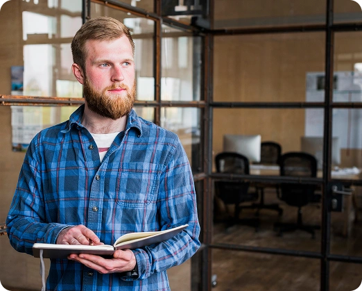 Op de foto houdt een man met een baard in een geruit overhemd een open notitieboekje vast en kijkt hij opzij. Hij staat in een moderne kantoorruimte met glazen scheidingswanden en een bureau achter zich.