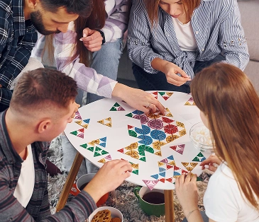 Een groep vrienden zit aan tafel en stelt samen een kleurrijk bordspel samen uit geometrische elementen. De sfeer is gezellig en vriendelijk: iedereen is bij het proces betrokken en gaat actief met elkaar om.