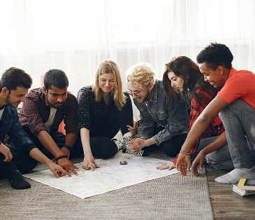 Een groep jonge vrienden zit op de grond en bestudeert aandachtig de grote kaart die voor hen ligt, wat erop wijst dat ze samen een reis of avontuur aan het plannen zijn.