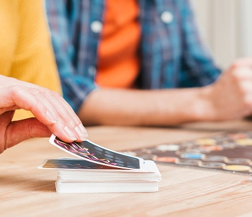 Twee mensen spelen een bordspel en een kaartspel aan een houten tafel; de ene trekt een kaart, terwijl de andere zijn telefoon vasthoudt.