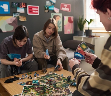 Op de foto zitten drie tieners aan een tafel en spelen ze een bordspel met kaarten, figuren en dobbelstenen. Een van hen houdt de kaarten in zijn handen, terwijl de anderen hun zetten overwegen.